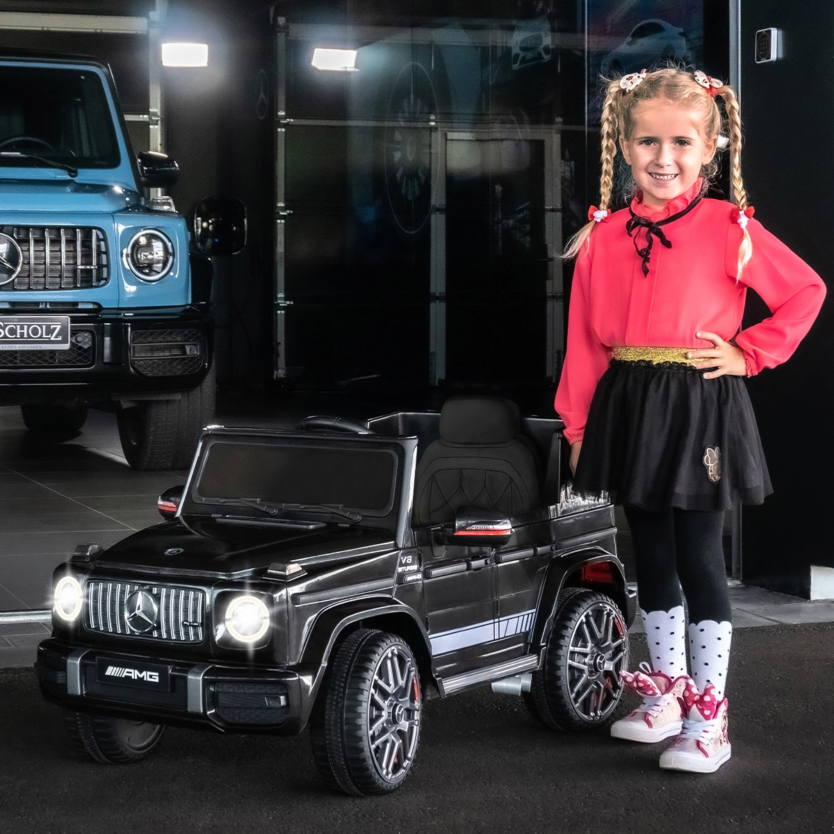 Actionbikes Kinder-Elektroauto Mercedes AMG G63 W463 Mädchen steht lächelnd neben dem Mercedes Kinderauto mit leuchtenden Scheinwerfern